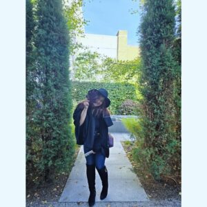 A woman wearing a black witch hat and cape stands in the garden at the Peabody Essex Museum in Salem, Massachusetts. She is playfully holding a dark-colored leaf in front of her face while smiling. The scene is framed by tall green trees and lush garden foliage, creating a serene and enchanting atmosphere.