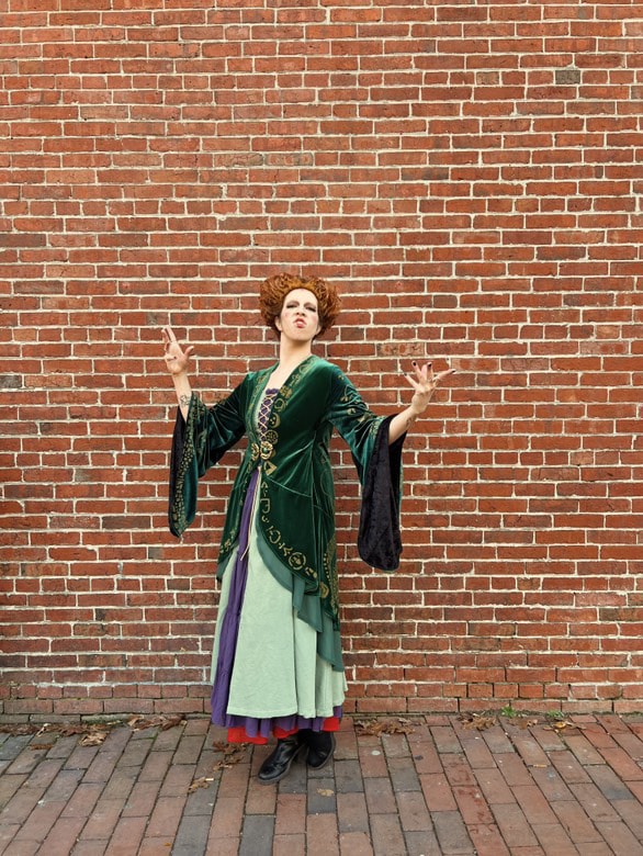 A person dressed as Winifred Sanderson from "Hocus Pocus" poses in front of a red brick wall in Salem, Massachusetts. They wear a green velvet robe with intricate designs and hold their hands in an expressive pose, giving a playful witchy vibe.