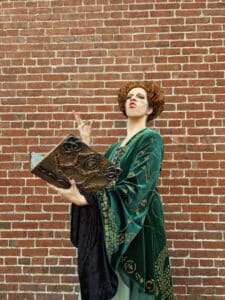 A person dressed as Winifred Sanderson from "Hocus Pocus" poses dramatically in front of a brick wall in Salem, Massachusetts. They are holding a spellbook and wearing a green velvet robe while making a casting gesture with their hand, looking witchy and confident.