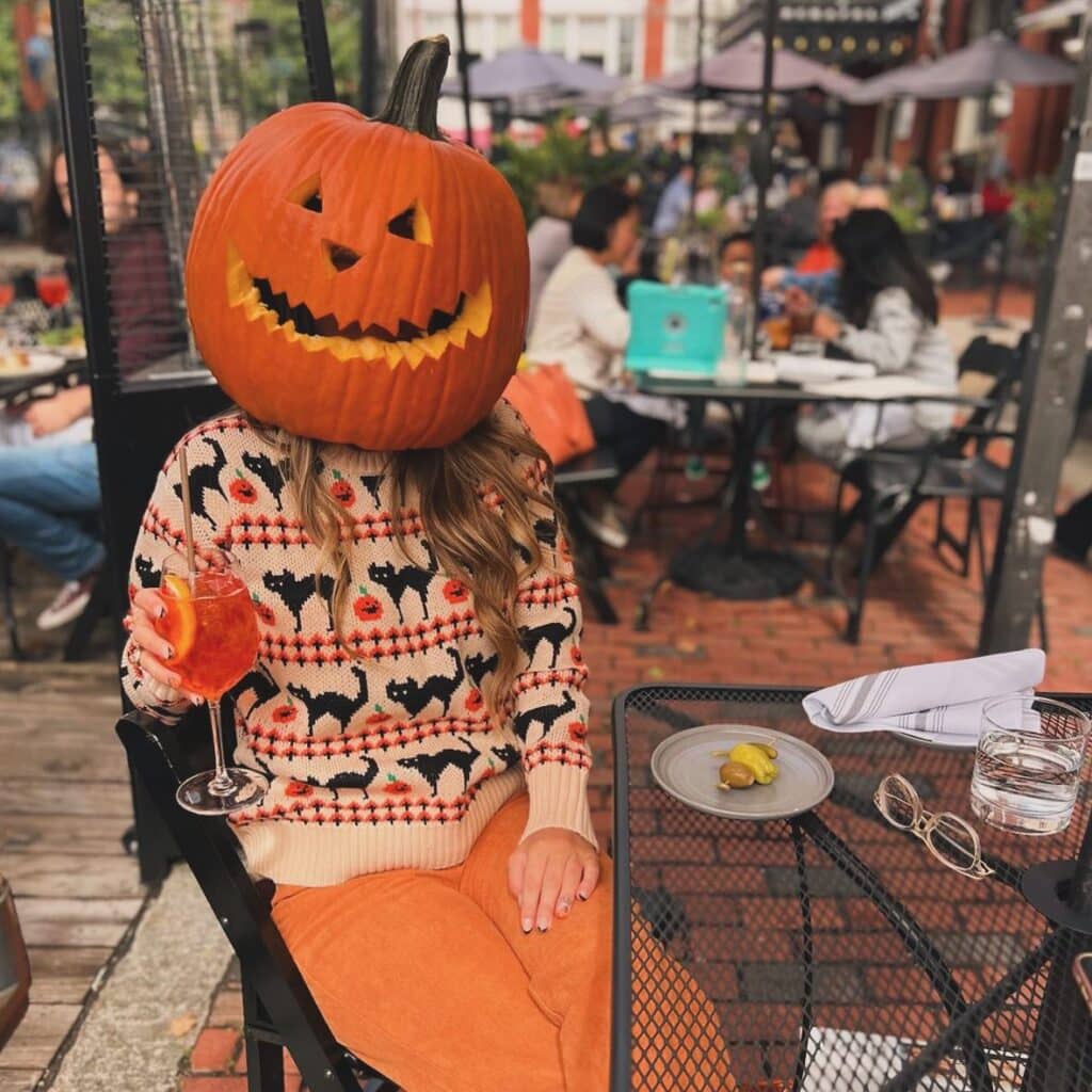 A playful Halloween scene at Ledger Restaurant in Salem, Massachusetts, with a diner wearing a carved pumpkin as a mask while enjoying a festive drink on the outdoor patio. Surrounded by fall vibes and a lively crowd, the moment captures the spirit of Salem’s famous Halloween season.