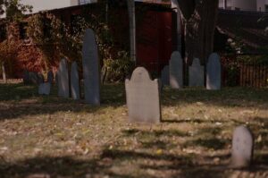A photo of the Old Burying Point Cemetery in Salem, Massachusetts, showing several old, weathered gravestones standing in a grassy area. The ground is covered with fallen leaves, and a large tree provides shade over the cemetery. In the background, a brick building is covered in ivy, adding to the peaceful and historic atmosphere.