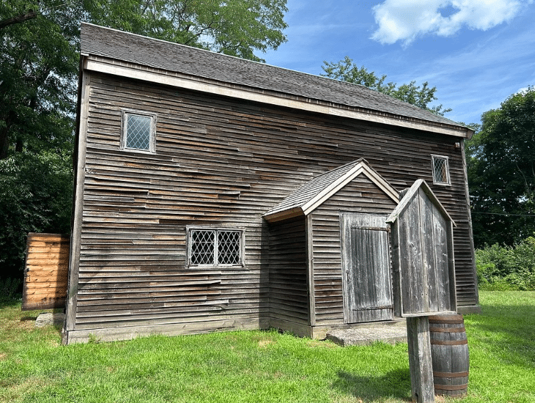 A historic wooden building at the Rebecca Nurse Homestead, featuring weathered clapboard siding, small diamond-paned windows, and a simple gabled entrance. The structure sits on a grassy lawn under a bright blue sky, with a wooden signboard and barrel in the foreground. This setting reflects the rustic charm and historical authenticity of the 17th-century homestead.