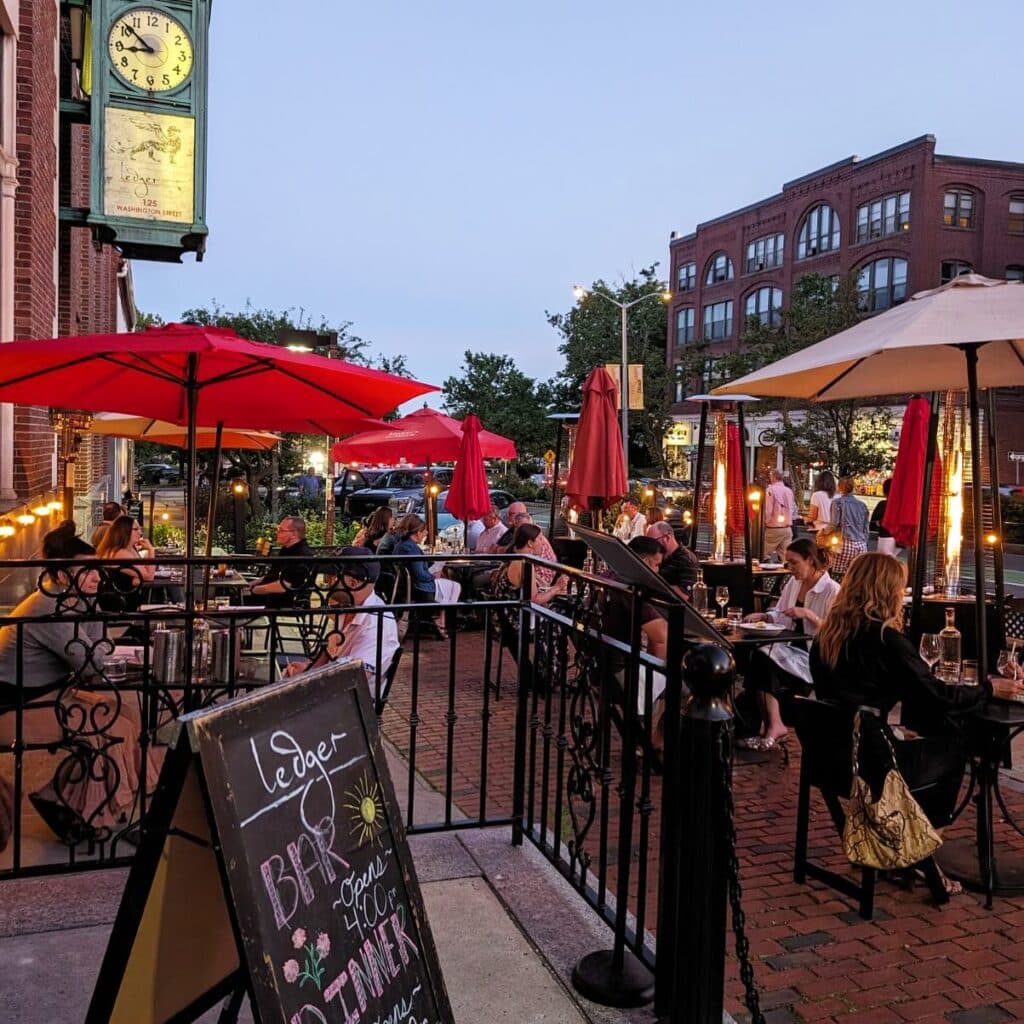 The outdoor patio at Ledger Restaurant in Salem, Massachusetts, offers a lively and welcoming atmosphere, with diners enjoying meals under red umbrellas and surrounded by warm evening lights. This charming space, set against the backdrop of historic Salem architecture, is perfect for casual dining or special occasions during the warmer months.