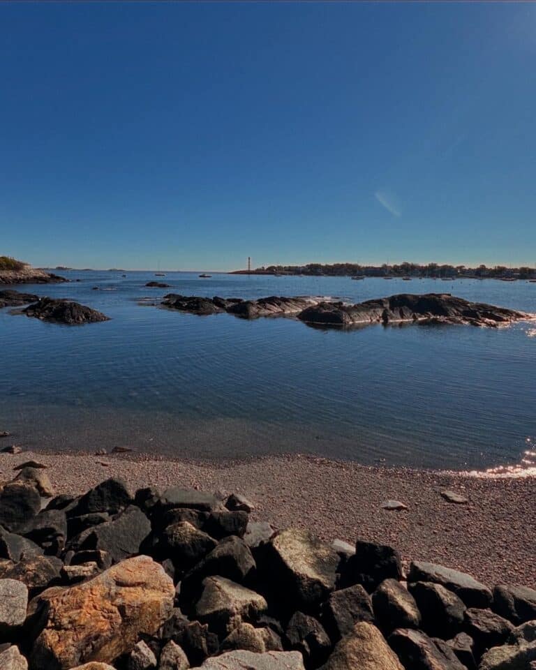 A photo of a rocky shoreline in Salem, Massachusetts, showing calm blue water and small rock formations scattered across the sea. The sky is clear and bright blue, and a distant lighthouse can be seen on the horizon, adding a touch of coastal charm.