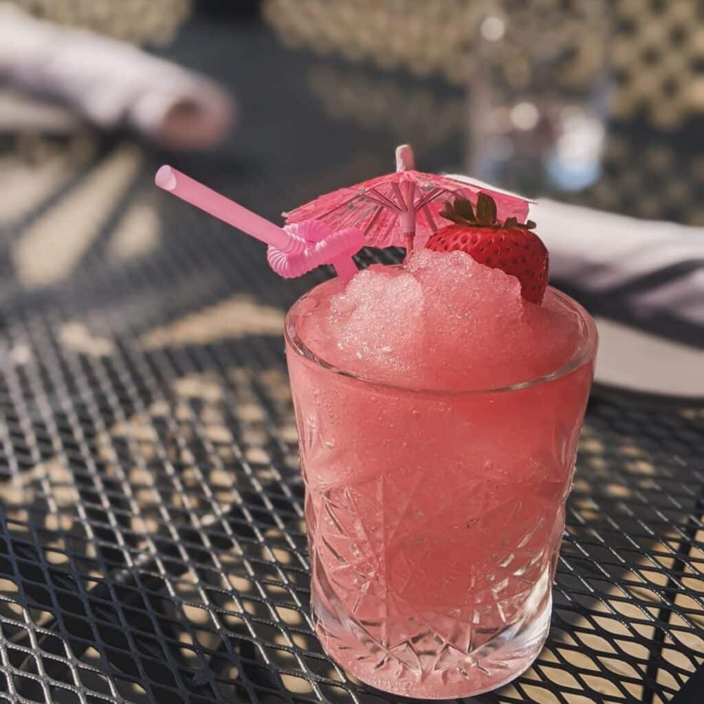 A refreshing frozen strawberry cocktail at Ledger Restaurant in Salem, Massachusetts, served in a textured glass with a fun pink umbrella, a straw, and a fresh strawberry garnish. Perfect for enjoying on a sunny day.