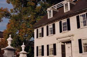 A photo of the Ropes Mansion in Salem, Massachusetts, showing its white exterior with black shutters and detailed trim. The house features dormer windows on the roof and a classic front entrance. In front of the mansion, two white stone pillars stand against a backdrop of lush green trees with hints of fall colors.