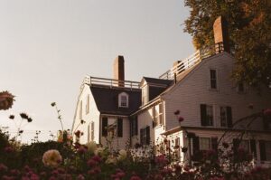 A photo of the Ropes Mansion in Salem, Massachusetts, with its white exterior, black shutters, and tall brick chimneys. The foreground features blooming flowers in shades of pink, white, and purple, creating a colorful and lively garden scene.