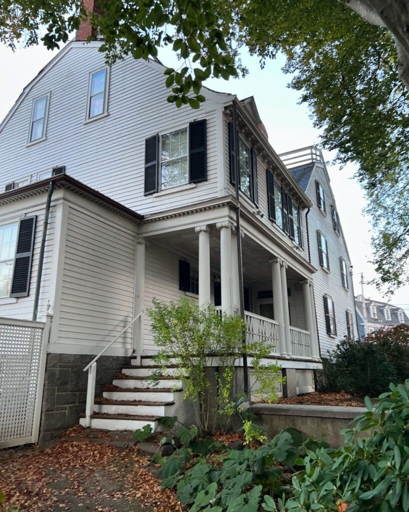 The Ropes Mansion in Salem, Massachusetts, a historic white Georgian Colonial home with black shutters and a front porch supported by elegant columns, surrounded by greenery and fallen autumn leaves. The house stands prominently under the shade of tall trees, exuding timeless charm.