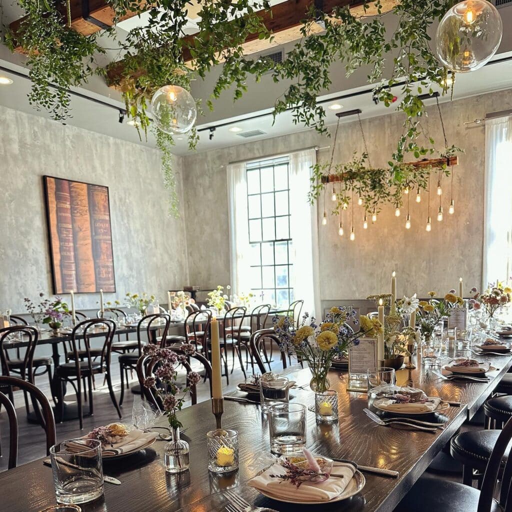 An elegantly arranged dining space at Ledger Restaurant in Salem, Massachusetts, featuring long wooden tables adorned with fresh floral centerpieces, glowing candles, and sophisticated place settings. The hanging greenery and warm Edison bulb lighting add a natural and inviting touch, creating the perfect atmosphere for private events, weddings, or special celebrations.