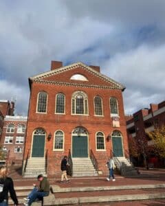 Salem Old Town Hall, a historic Federal-style brick building with green doors and large arched windows, stands prominently under a cloudy sky in Salem, Massachusetts. People walk along the brick-paved square in front of the building, adding a lively touch to the historic scene.