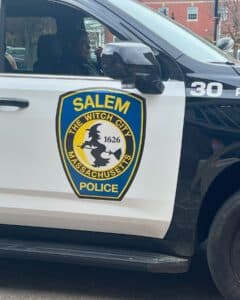 A Salem Police cruiser in Salem, Massachusetts, displaying the "Witch City" emblem on its door, featuring a silhouette of a witch on a broomstick and the year "1626." A police officer is visible inside the vehicle, with brick buildings in the background.