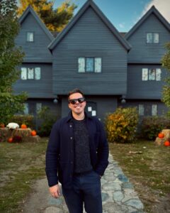 A person wearing sunglasses and a dark outfit standing on a stone path in front of the historic Witch House in Salem, Massachusetts, a dark timber-framed building with three gabled roofs, surrounded by autumn decorations with pumpkins and hay bales.