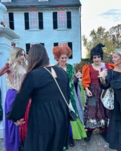 A group of people dressed as the Sanderson Sisters from Hocus Pocus pose in front of the historic Ropes Mansion in Salem, Massachusetts. The white colonial house with black shutters and glowing red-lit windows serves as the perfect backdrop for the festive Halloween costumes.