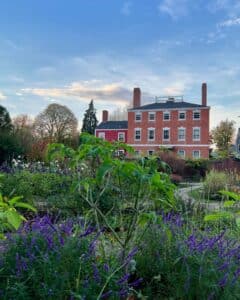 The Ropes Mansion Garden in Salem, Massachusetts, featuring vibrant purple flowers and lush greenery in the foreground, with the historic red brick Ropes Mansion standing prominently under a pastel blue sky in the background. The peaceful setting is framed by trees and pathways winding through the garden.