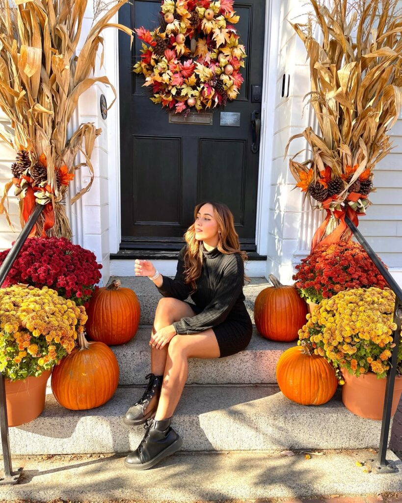 A woman sitting on the steps of a charming home in Salem, Massachusetts, surrounded by fall decorations, including pumpkins, colorful mums, cornstalks, and a vibrant autumn wreath.