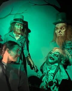 A creepy leprechaun-themed display in Salem features eerie figures with green-tinted lighting, including two tall, ghastly characters in top hats and a smaller, grinning figure with sharp teeth and wild hair. A visitor glances at the scene, adding a sense of curiosity and amusement to the spooky setup.
