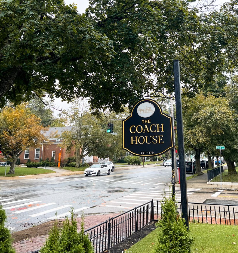 A view of The Coach House sign on a rainy day in Salem, Massachusetts. The black and gold sign reads "The Coach House, Est. 1879" and features an illustration of a horse-drawn carriage, hinting at the inn’s historic roots. The sign stands beside a quiet intersection with trees lining the street and a few cars passing by. The greenery, iron fence, and trees give the scene a cozy, timeless atmosphere, inviting visitors to experience the charm of this historic boutique inn.