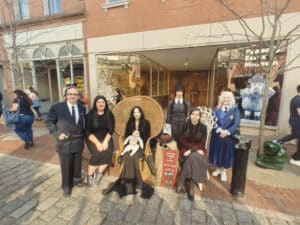 A group of people dressed as characters from the Addams Family pose on a sidewalk in Salem, Massachusetts. They are in front of the Peabody Essex Museum, with costumes representing Gomez, Morticia, Wednesday, Pugsley, and other iconic characters.