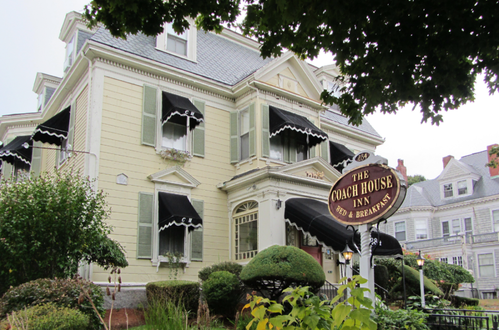 The Coach House Inn, a charming bed and breakfast in Salem, Massachusetts, with a pale yellow exterior and classic New England architectural details. The building features green shutters, black awnings with scalloped edges, and a decorative porch entrance. A vintage-style sign in the foreground reads "The Coach House Inn Bed & Breakfast" and displays the address "284." Manicured bushes and greenery surround the inn, while neighboring historic homes add to the picturesque setting. The scene is inviting, blending historic elegance with a cozy, welcoming atmosphere.