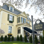A charming historic inn with a pale yellow exterior and green shutters, The Coach House stands under a clear sky. The entrance features a black awning, and the manicured lawn is lined with tall shrubs. A black and gold sign reading "The Coach House, Est. 1879" is visible in the foreground, giving a welcoming touch to this elegant New England-style building. Branches from a nearby tree frame the top of the image.