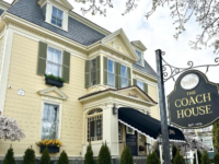 A charming historic inn with a pale yellow exterior and green shutters, The Coach House stands under a clear sky. The entrance features a black awning, and the manicured lawn is lined with tall shrubs. A black and gold sign reading "The Coach House, Est. 1879" is visible in the foreground, giving a welcoming touch to this elegant New England-style building. Branches from a nearby tree frame the top of the image.