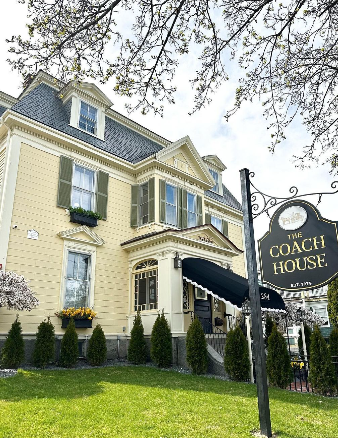 A charming historic inn with a pale yellow exterior and green shutters, The Coach House stands under a clear sky. The entrance features a black awning, and the manicured lawn is lined with tall shrubs. A black and gold sign reading "The Coach House, Est. 1879" is visible in the foreground, giving a welcoming touch to this elegant New England-style building. Branches from a nearby tree frame the top of the image.