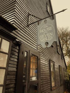 A close-up photo of The House of the Seven Gables in Salem, Massachusetts, showing its dark wooden siding and the sign hanging above the entrance. The sign reads "The House of the Seven Gables" in elegant lettering, while the surrounding building has a rustic, historic charm.