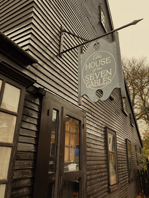 A close-up photo of The House of the Seven Gables in Salem, Massachusetts, showing its dark wooden siding and the sign hanging above the entrance. The sign reads "The House of the Seven Gables" in elegant lettering, while the surrounding building has a rustic, historic charm.