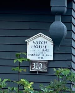 A close-up of the Witch House sign in Salem, Massachusetts, featuring a small white plaque with the text "Witch House, Built ca. 1642, Restored by Historic Salem Inc. 1946" and the house number "310" below it. The dark wooden exterior of the house and surrounding greenery provide a striking backdrop.
