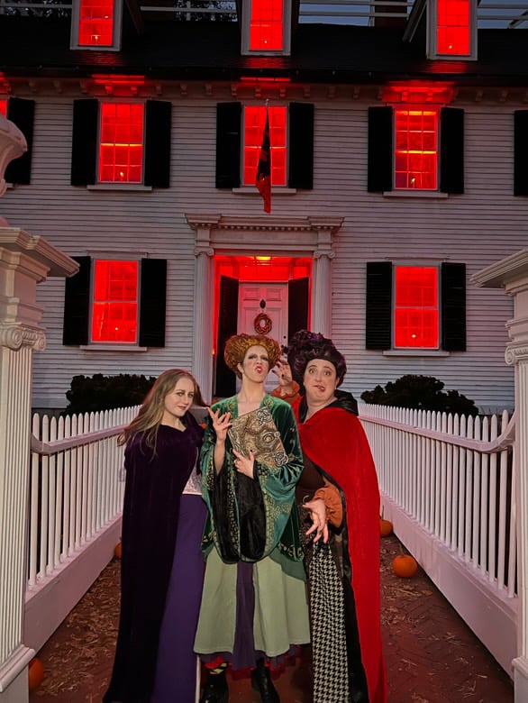 Three people dressed as the Sanderson Sisters from "Hocus Pocus" are standing in front of the Ropes Mansion in Salem, Massachusetts, illuminated by glowing red lights in the windows, enhancing the eerie atmosphere.