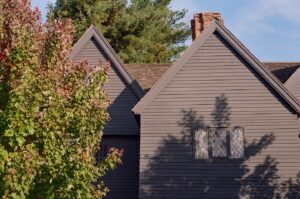 A photo of the Witch House in Salem, Massachusetts, showing its dark wooden exterior with steep gabled roofs and diamond-paned windows. A large green tree partially obscures the left side of the house, while the sunlight casts shadows across the walls, adding to the historic atmosphere