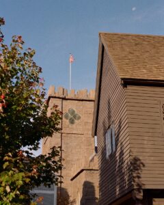 A photo showing the Witch House in Salem, Massachusetts, with its dark wooden exterior and steep roof. In the background, the stone tower of the First Church of Salem rises with an American flag on top, framed by a clear blue sky and partially hidden behind green trees.