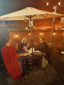 Three women dressed as the Sanderson Sisters from Hocus Pocus sitting at an outdoor cafe table under an umbrella near Salem Common, with soft evening lighting and a brick wall decorated with hanging string lights in the background.
