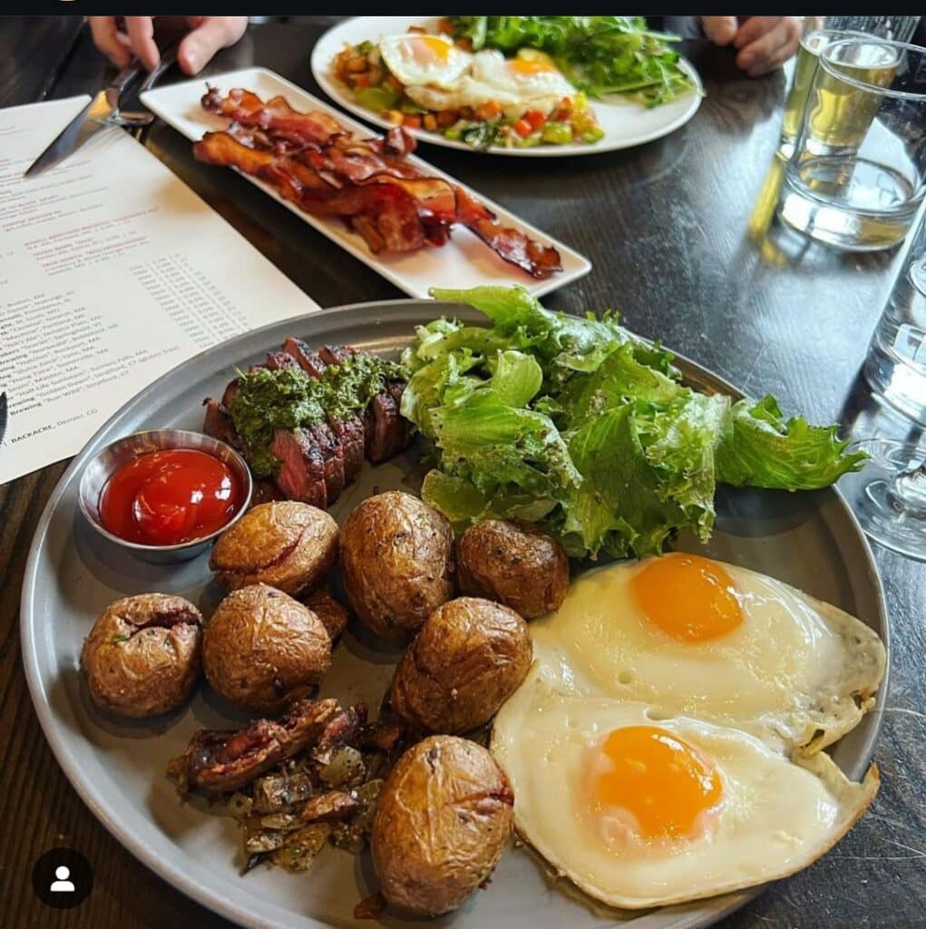 A beautifully plated brunch at Ledger Restaurant in Salem, Massachusetts, featuring crispy roasted potatoes, sunny-side-up eggs, a fresh green salad, and sliced steak topped with chimichurri sauce. In the background, a plate of crispy bacon and another dish with eggs and vegetables complete the delicious spread.