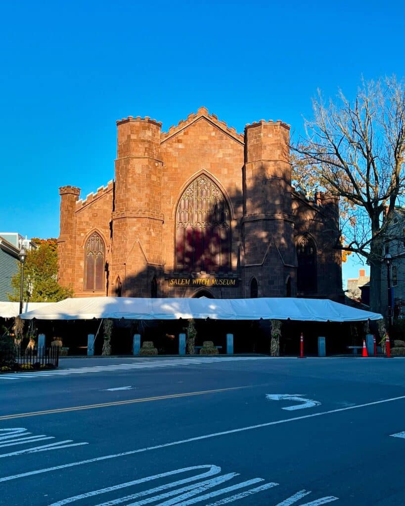 A photograph of the Salem Witch Museum, housed in a striking Gothic-style building made of red stone. The large arched windows and imposing architecture give it a historic and dramatic appearance, fitting for its focus on the events of the 1692 Salem Witch Trials. A white tent is set up outside the museum, surrounded by seasonal decorations such as hay bales and cornstalks, adding a festive autumn atmosphere. The bright blue sky and bare tree branches provide a seasonal contrast, enhancing the museum's allure as a must-visit destination for those interested in the history of the Salem Witch Trials.