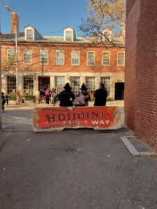 A view of three people sitting behind a concrete barrier painted with the words "Houdini Way" in Salem, Massachusetts. The scene includes two people dressed as witches, adding to the Halloween atmosphere. In the background, there's a red brick building with large windows and people walking by on the street.
