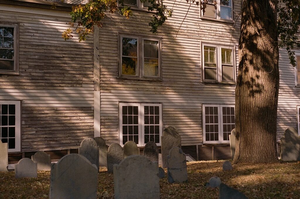 A photo of the Old Burying Point Cemetery in Salem, Massachusetts, showing several aged gravestones in front of a weathered wooden house. The house has old, faded siding with multiple windows, and a large tree casts dappled shadows across both the building and the gravestones. The sunlight filters through the leaves, creating a calm and historic atmosphere.