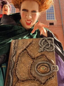 A close-up of a girl dressed as Winifred Sanderson from the movie Hocus Pocus in Salem, Massachusetts. She holds a large spellbook with an eye on the cover and snake decorations. Her bright red wig and green robe add to her mischievous expression.