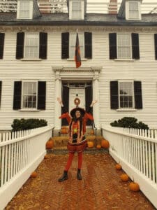 A woman dressed in a festive witch costume stands at the entrance of the Ropes Mansion in Salem, Massachusetts, smiling with her arms raised. Pumpkins decorate the pathway leading to the mansion's white facade, with autumn leaves scattered around.
