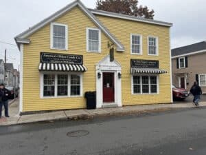 A yellow clapboard building with white trim and a red front door, home to Ye Olde Pepper Companie, America’s oldest candy company, located in Salem, Massachusetts. The building features two black-and-white striped awnings with signs reading "America's Oldest Candy Co." and "Ye Olde Pepper Companie Manufacturing Confectioner Since 1806." The street in front is wet, suggesting recent rain, and a few pedestrians are visible nearby.