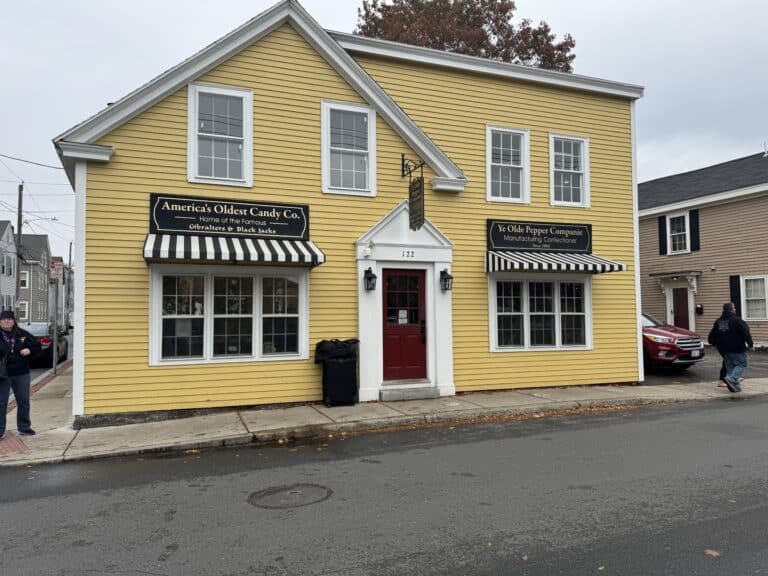 A yellow clapboard building with white trim and a red front door, home to Ye Olde Pepper Companie, America’s oldest candy company, located in Salem, Massachusetts. The building features two black-and-white striped awnings with signs reading "America's Oldest Candy Co." and "Ye Olde Pepper Companie Manufacturing Confectioner Since 1806." The street in front is wet, suggesting recent rain, and a few pedestrians are visible nearby.