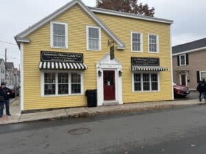 The storefront of Ye Olde Pepper Companie, a yellow wooden building with white trim and a red door, located in Salem, Massachusetts. The building features two striped black-and-white awnings with signs above them reading "America's Oldest Candy Co." and "Ye Olde Pepper Companie Manufacturing Confectioner Since 1806." The wet pavement suggests recent rain, with a few pedestrians visible near the building.