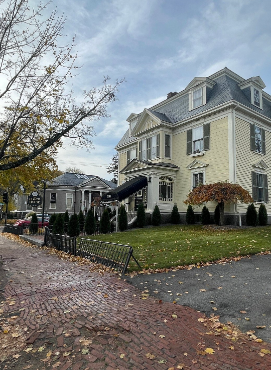 The Coach House Inn in Salem, Massachusetts, a historic yellow building with classic New England architectural details. The inn features green shutters, a black awning over the entrance with the number "284," and a neatly landscaped lawn with hedges and a small tree showing autumn colors. A cobblestone sidewalk and fallen leaves add a seasonal charm to the scene, and a sign reading "The Coach House Inn" is visible by the roadside. The sky is lightly overcast, contributing to a cozy, inviting atmosphere in this picturesque Salem neighborhood.
