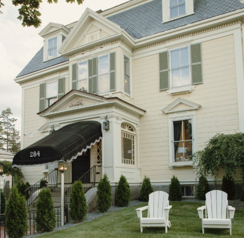 A historic yellow inn with green shutters, The Coach House features a black awning over the entrance displaying the address "284." The building’s classic New England architecture includes detailed trim and large windows. In front of the inn, two white Adirondack chairs sit on a well-maintained lawn, framed by neatly trimmed shrubs. The scene gives a welcoming and peaceful vibe, perfect for relaxing outdoors.