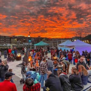 Crowds gathered at the Salem Massachusetts Frozen Fire Festival beneath a glowing, fiery sunset, enjoying warm fire pits, festive treats, and a joyful winter gathering.