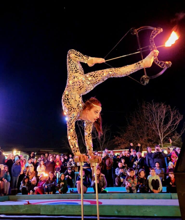 A skilled performer in Salem Massachusetts balancing upside down on hand supports, drawing a bow with her foot and aiming a flaming arrow, captivating the crowd at the Frozen Fire Festival.