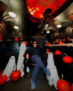 A person sitting on a black throne surrounded by small ghost figures and pumpkins at the Blackcraft store in Salem, with a red-lit ceiling and witch decoration in the background.
