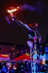 A skilled acrobat balances on her hands while using her feet to aim a flaming arrow from a bow during a nighttime performance in Salem, Massachusetts.