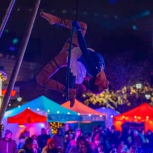 An aerial performer in a blue and white costume captivates the crowd at the Salem Frozen Fire Festival, with colorful tents and twinkling lights in Salem, Massachusetts.