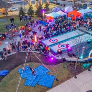 A vibrant aerial view of the Salem Frozen Fire Festival in Salem Massachusetts, featuring curling lanes, illuminated tents, and a crowd enjoying live performances and activities.